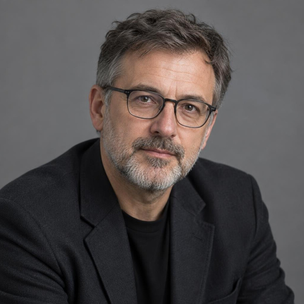 Studio portrait of a greying bearded man with glasses in a dark blazer